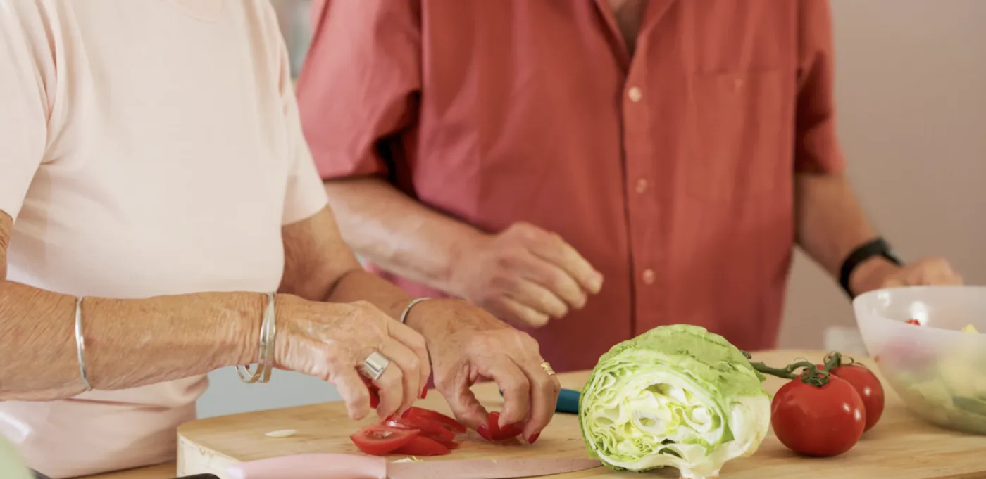 Two older people preparing a fresh salad together in the kitchen, slicing tomatoes on a wooden board